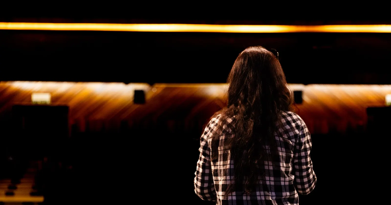 Una giovane donna di spalle, in camicia a quadri, sola sul palco davanti a una platea vuota dalle luci aranciate