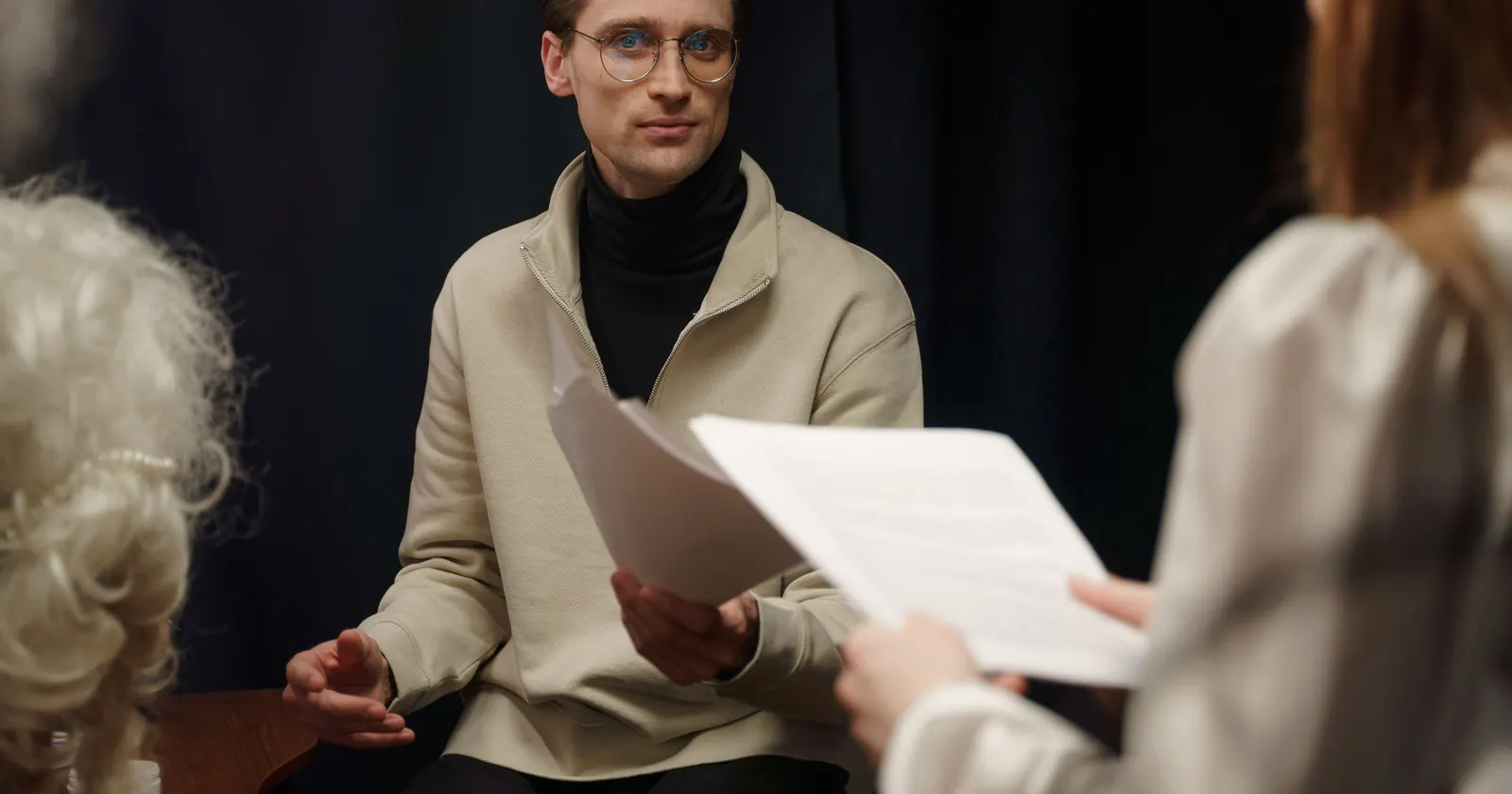 An actor listens to the person reading opposite him, seen from behind in the foreground with the script in hand