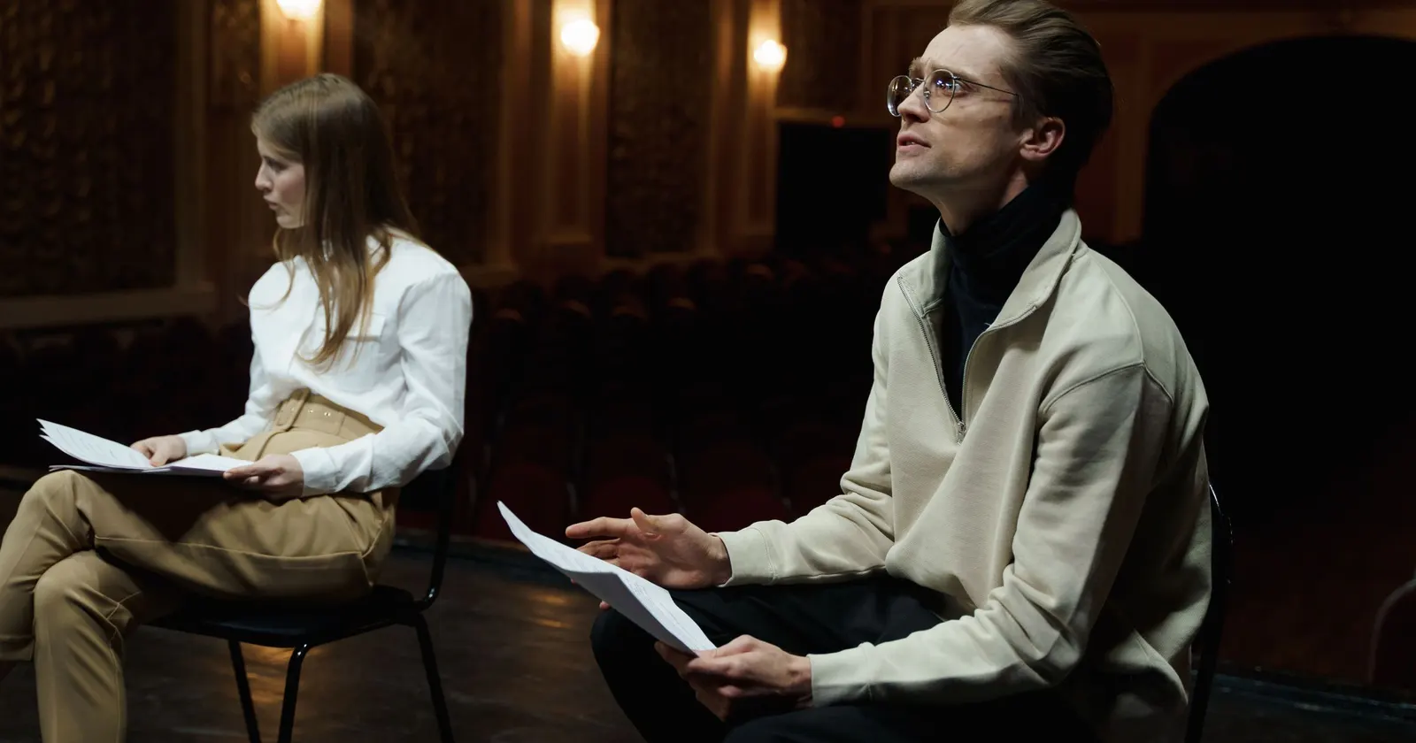 Deux comédiens assis sur une scène de théâtre, leur texte à la main ; lui lève les yeux, comme pour retrouver sa réplique